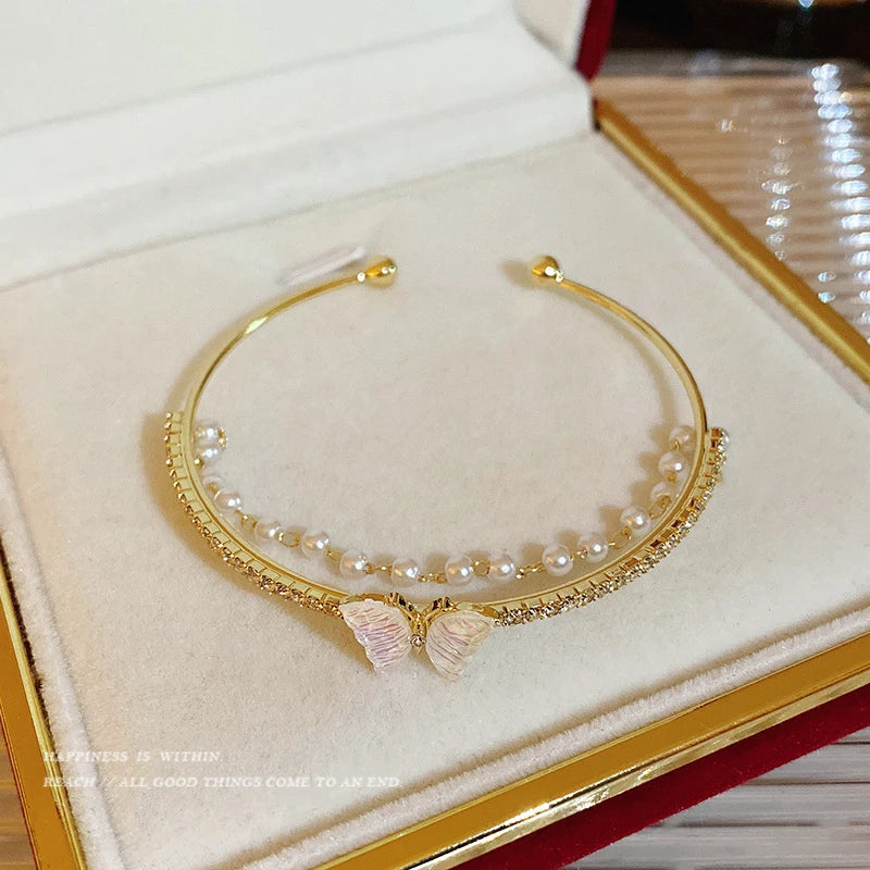 Pulseira "Aurum Elegance Butterfly" – Um acessório delicado e sofisticado com detalhes em pérolas e zircônias brilhantes, complementado por um gracioso detalhe de borboleta esmaltada em tons suaves. O design aberto facilita o uso e ajusta-se perfeitamente ao pulso, sendo ideal para ocasiões especiais ou para adicionar um toque de feminilidade ao dia a dia.